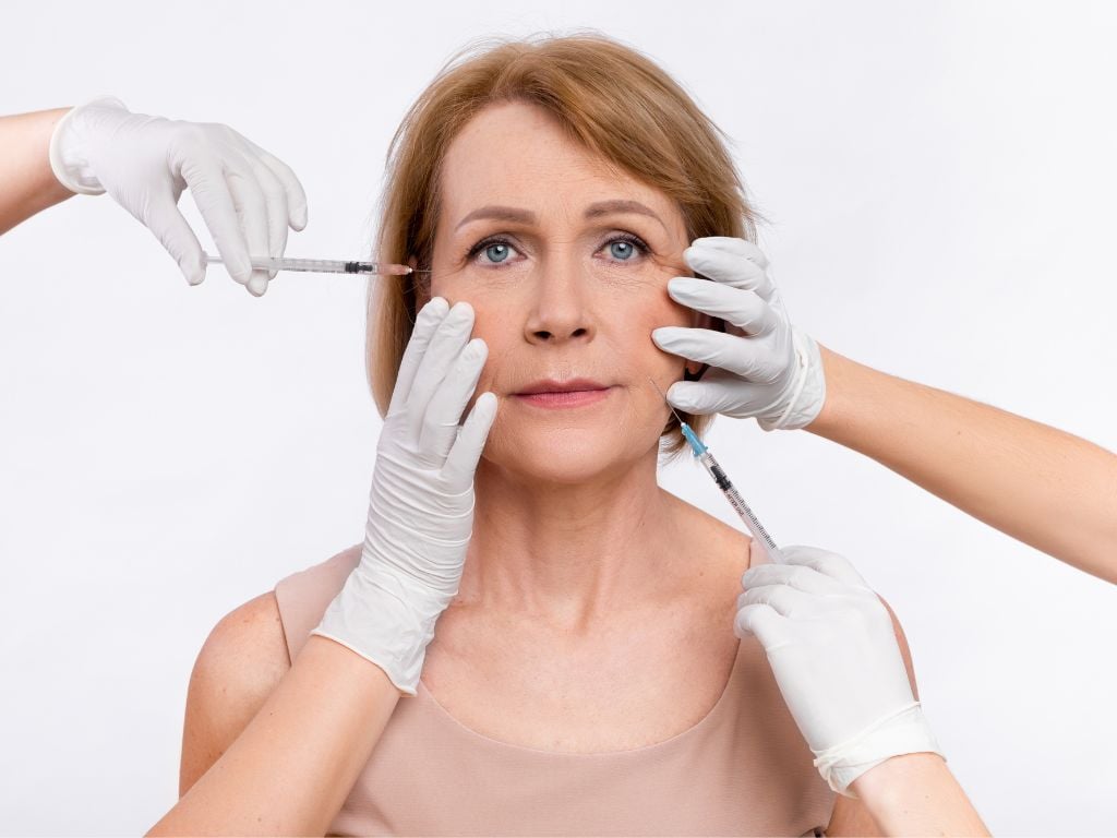 Mature woman surrounded by gloved hands holding syringes