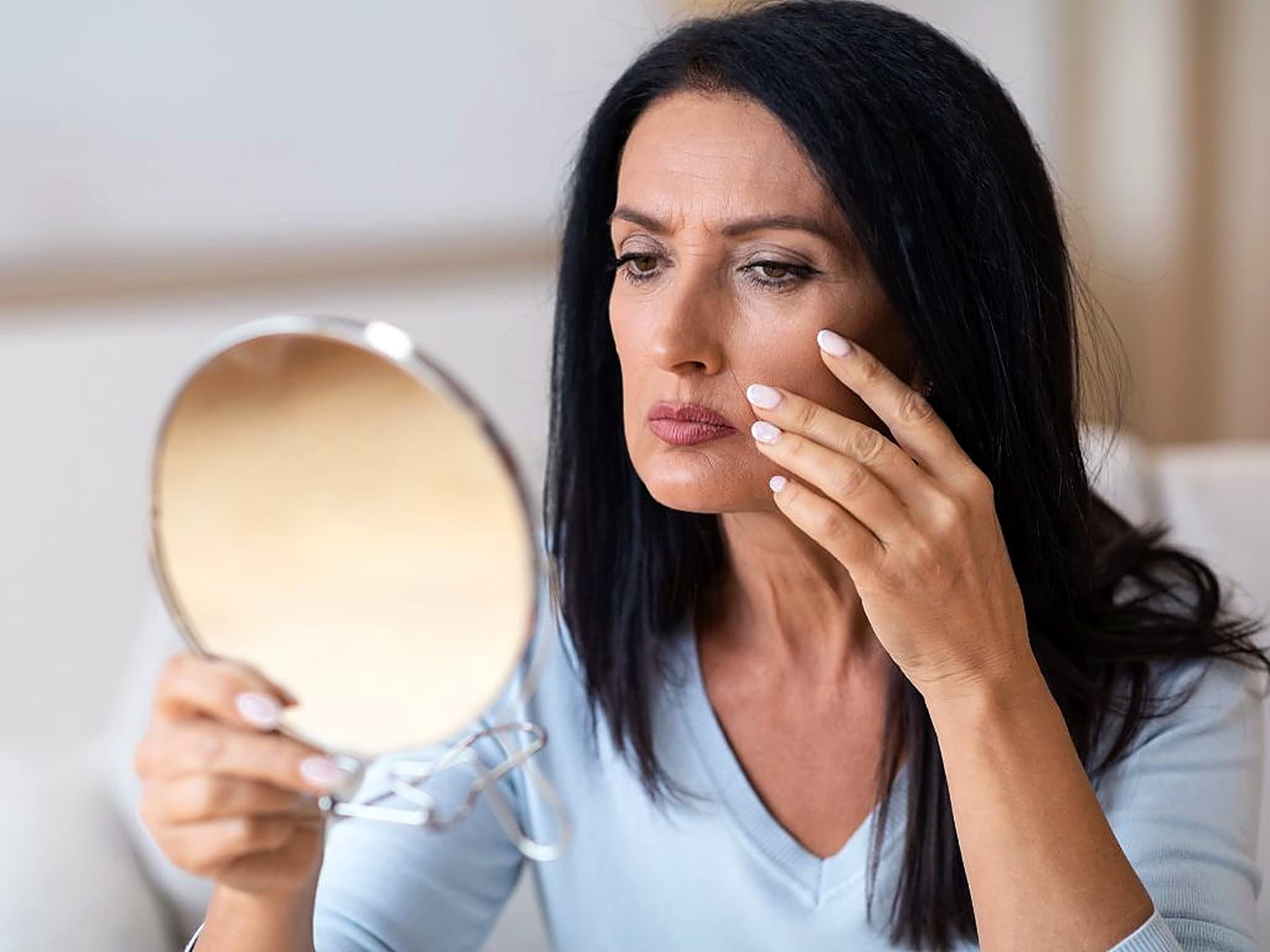 Woman looking into a hand mirror, examining the fine lines under her eyes