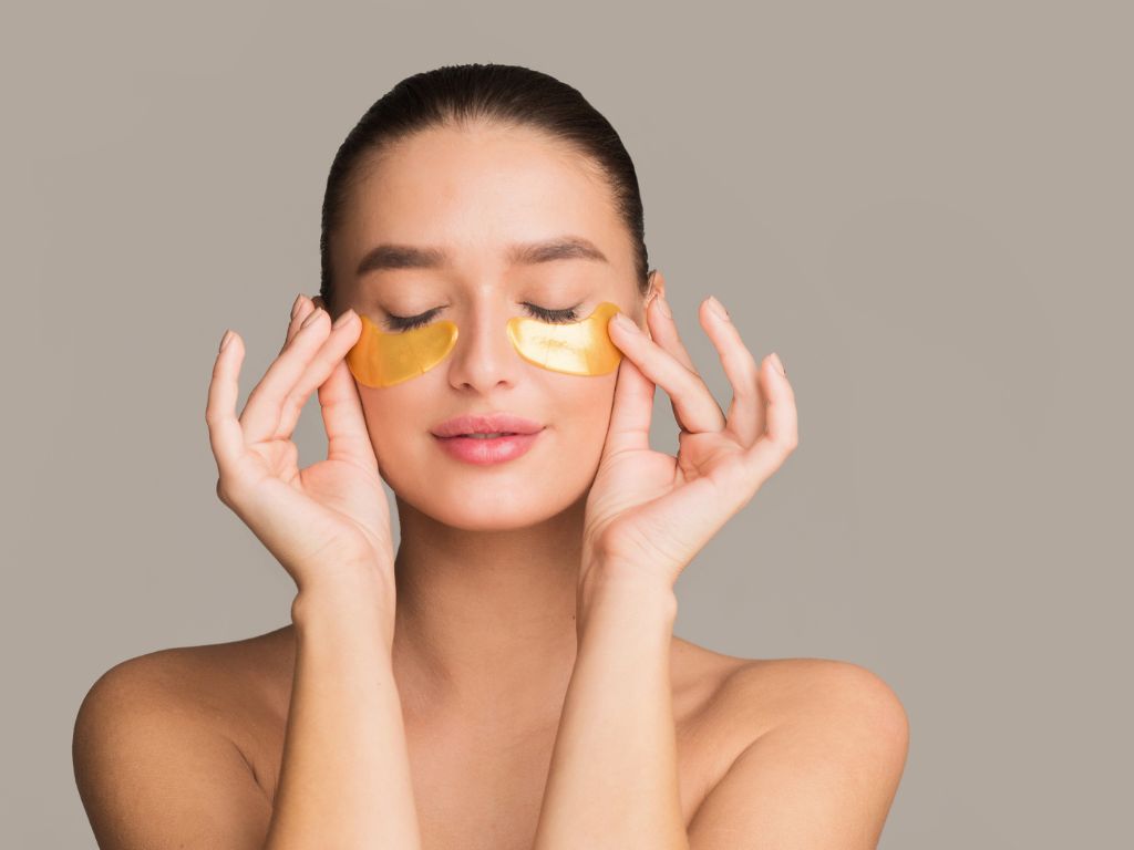 Γυναίκα με κλειστά μάτια εφαρμόζει χρυσά eye patches στην περιοχή κάτω από τα μάτια.